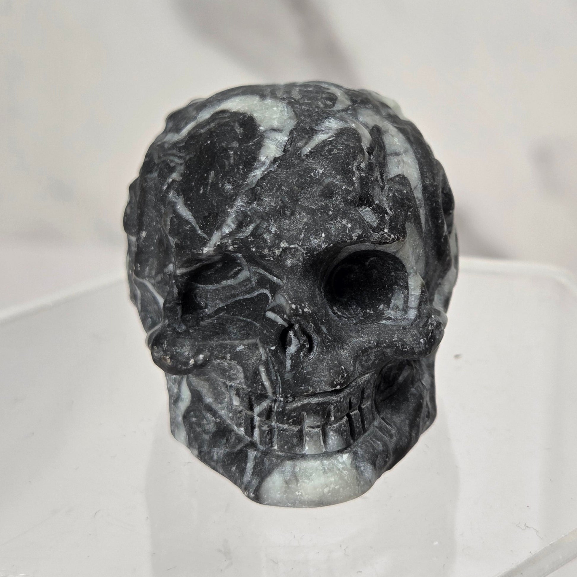 A carved black skull figurine with natural gray and white marbled patterns, displayed on a clear acrylic stand against a light gray marble-patterned background.