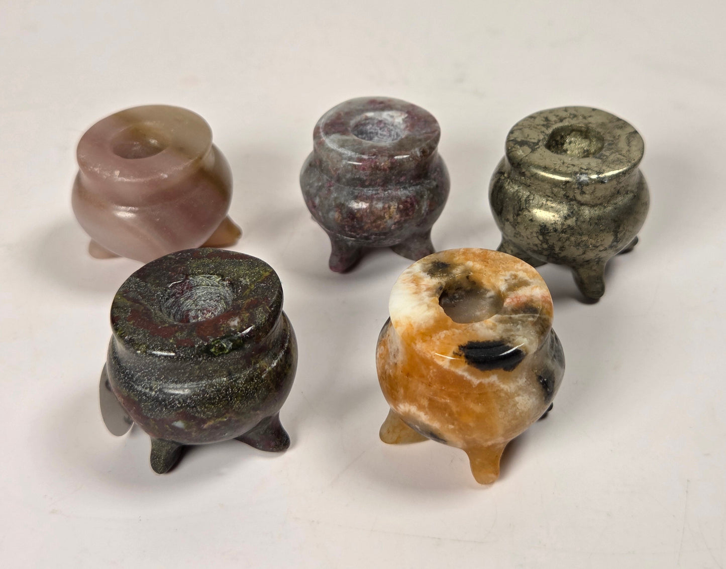 Five small carved stone censer-like objects with rounded bodies and short legs, arranged on a plain light background. The stones show varied natural colors including pink, reddish-brown, gray-green, dark charcoal, and orange-brown with dark spots.
