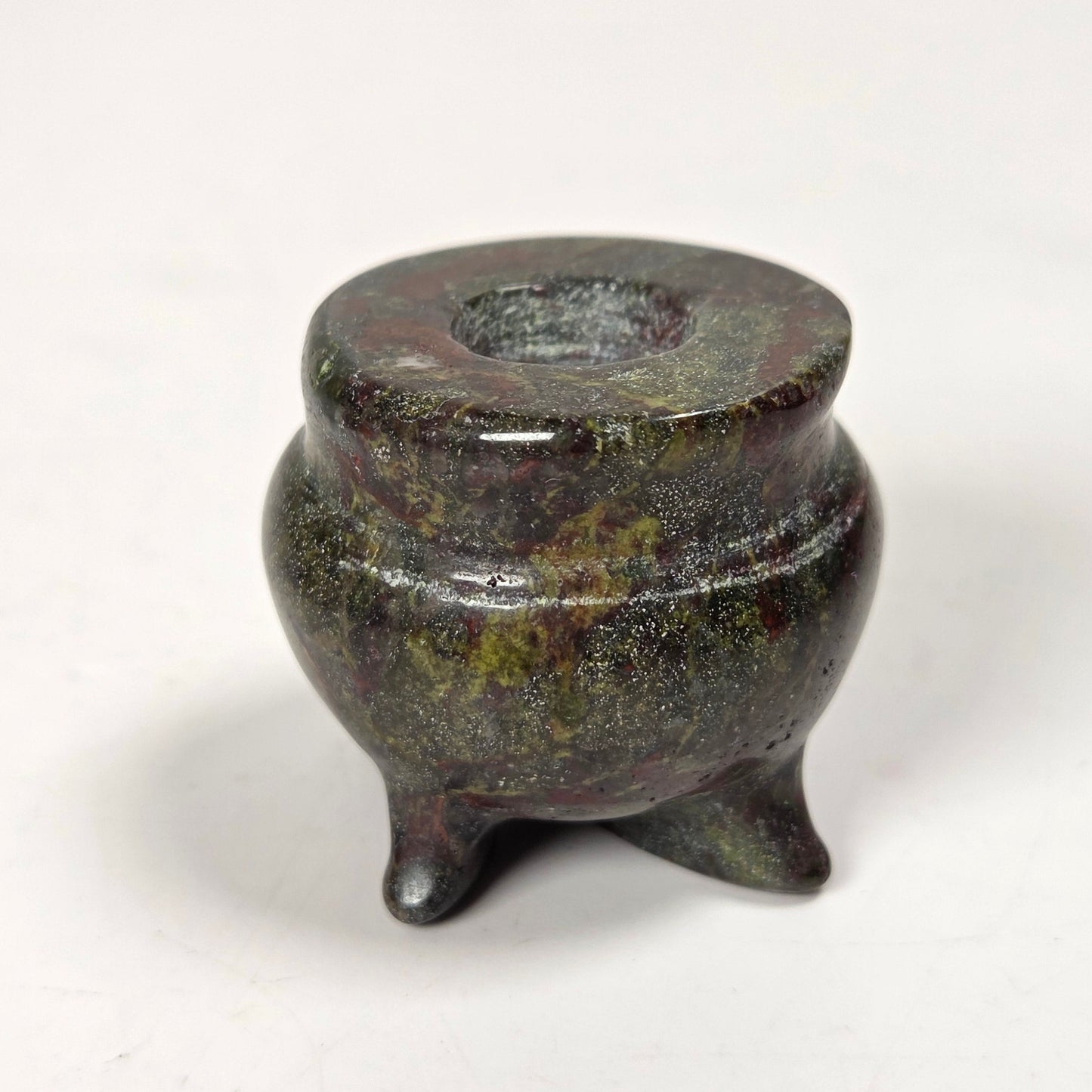 A small three-legged tripod vessel or candle holder made of dark polished stone or metal with a mottled green and bronze finish, set against a plain white background.