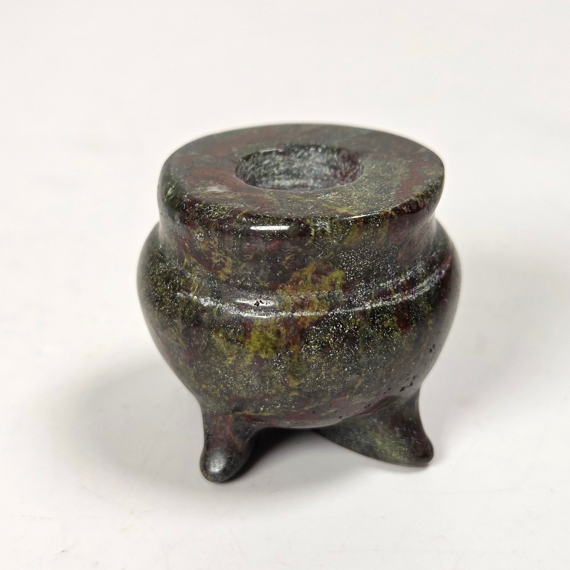 A small three-legged tripod vessel or candle holder made of dark polished stone or metal with a mottled green and bronze finish, set against a plain white background.