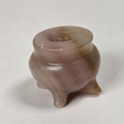 A small three-legged ceramic pot or candelabra made of smooth, polished stone with a mottled pinkish-brown and gray coloration, set against a plain off-white background.