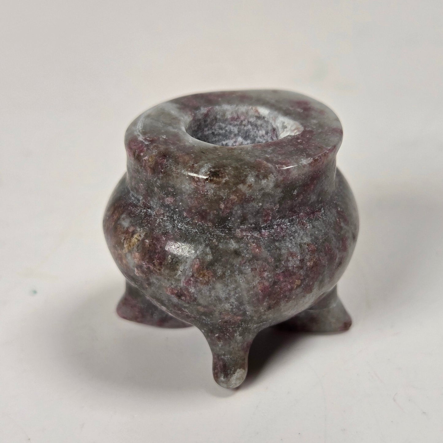 A small three-legged ceramic pot or candelabra made of smooth, polished stone with a mottled pinkish-brown and gray coloration, set against a plain off-white background.