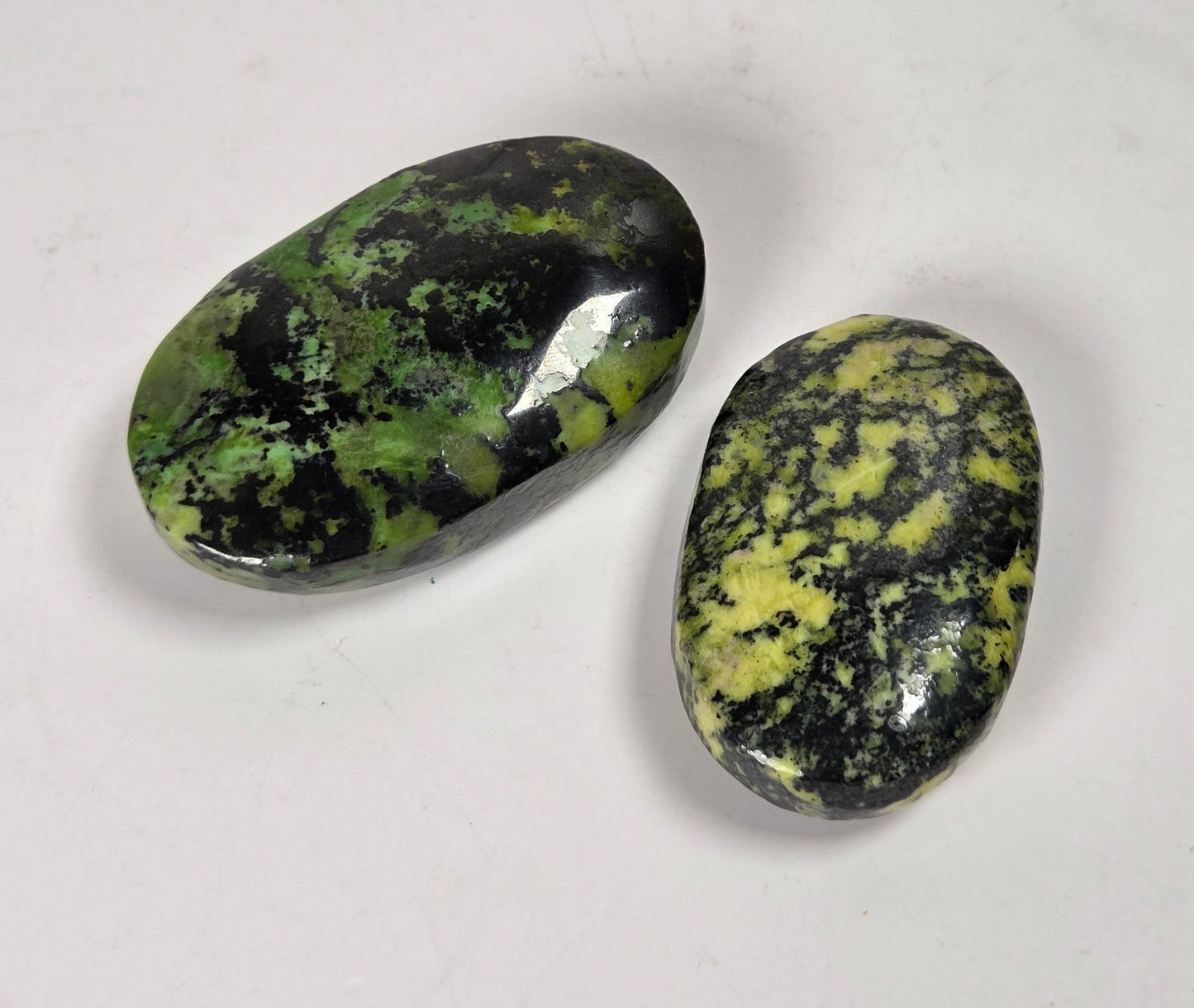 Two polished tumbled stones with a green and black mottled pattern, displayed on a plain light gray background.