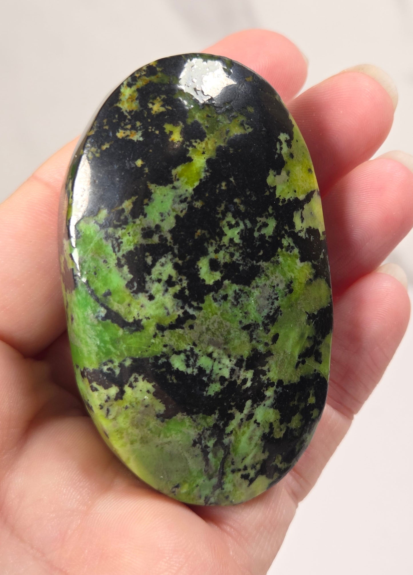 A hand holding a polished oval stone with a mottled pattern of black and green, photographed against a light marble-like background.