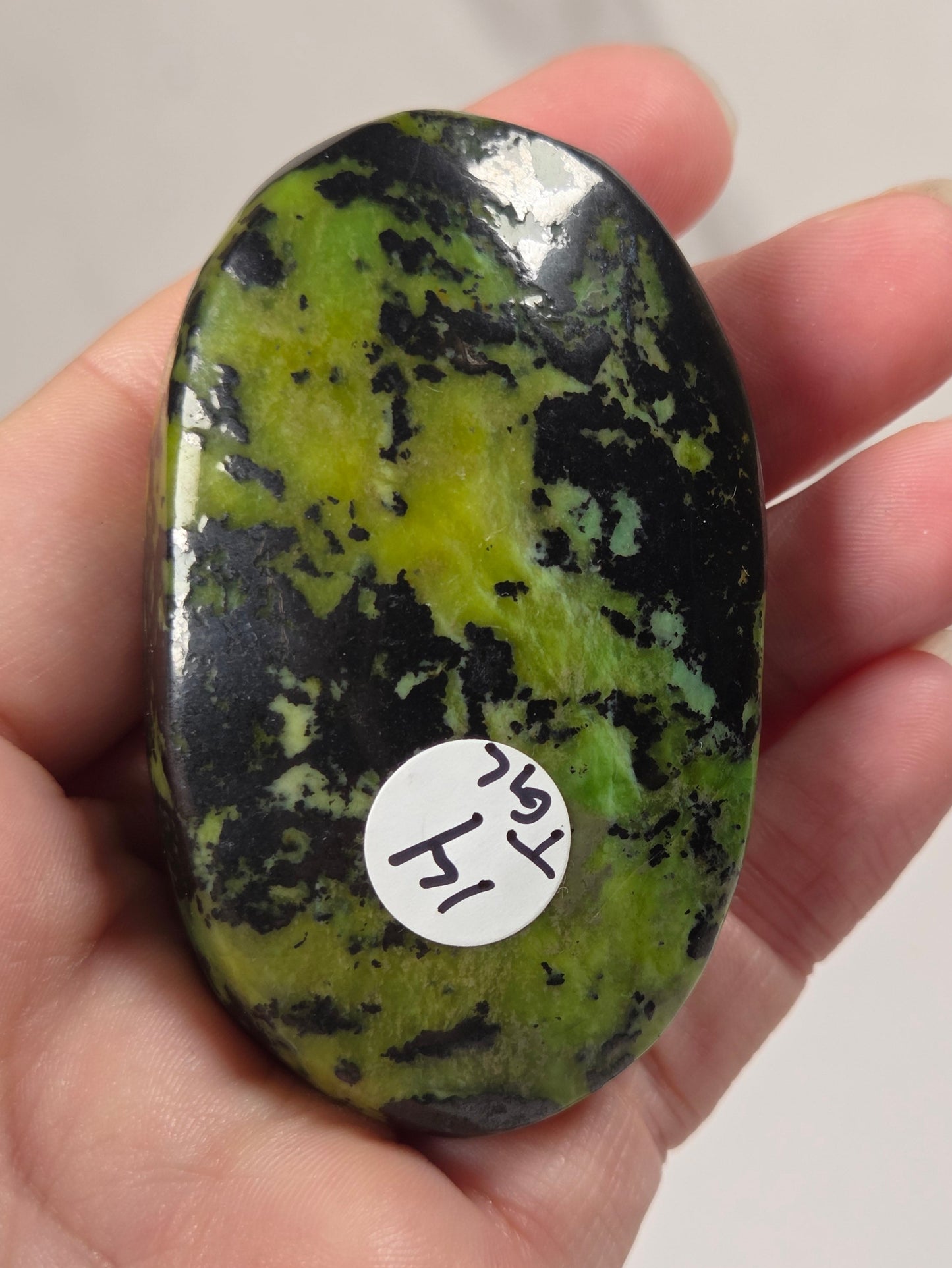 A hand holding a polished, oval-shaped green and black stone with a small white sticker on it, set against a light gray marble background.