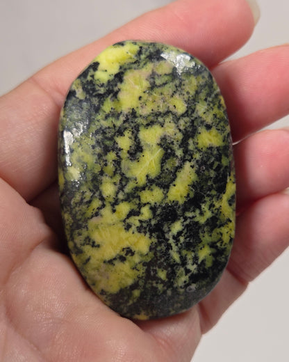 A hand holding a polished, oval-shaped stone with a marbled pattern of bright yellow and dark green.