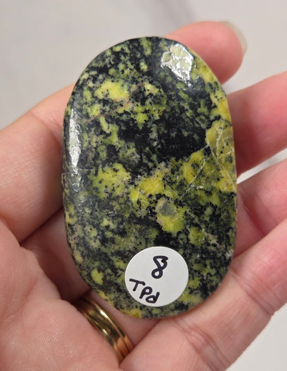 A hand holds a polished, oval-shaped green and black stone with bright yellow-green mottled patches. A small white circular sticker with the text '8 RPL' is attached to the bottom of the stone.