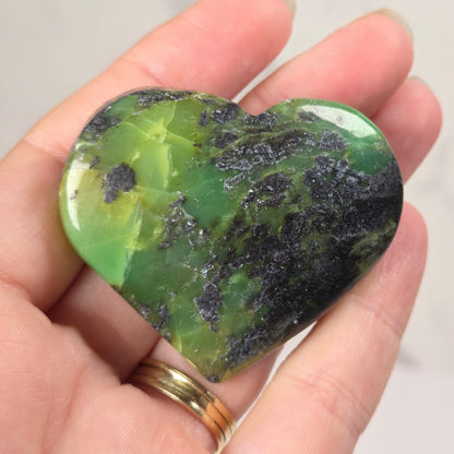 A heart-shaped polished green stone with dark natural inclusions, held in a person's hand against a light marble-like background.