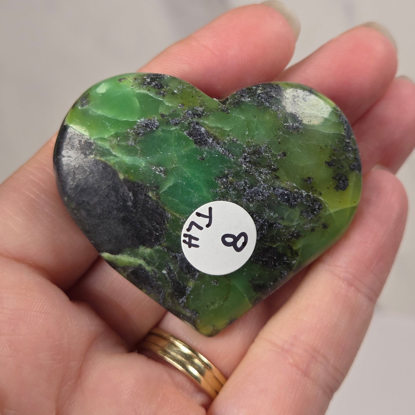A heart-shaped green stone with black veining, held in a person's hand. A small white circular sticker with 'HJ 8' is attached to the stone.