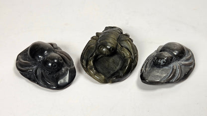 Three carved stone figurines shaped like spiders and scoprions, displayed side by side on a plain off-white surface. The left and right figurines are polished black, while the center cat figurine is carved from dark gray stone with natural veining.