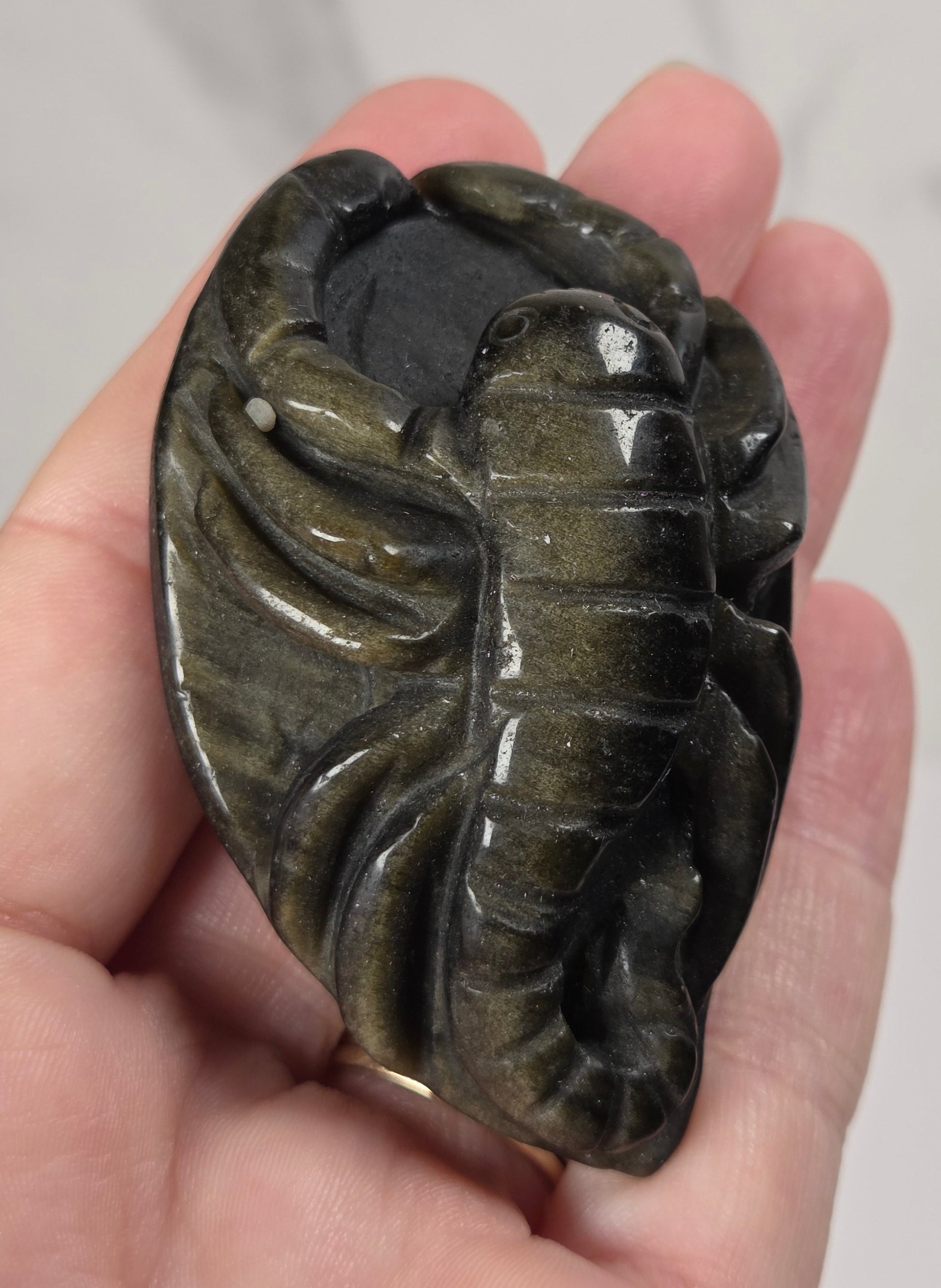 A hand holding a polished carved stone pendant shaped like a lobster, showing a dark olive-green and black marbled pattern.