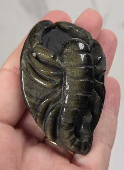 A hand holding a polished carved stone pendant shaped like a lobster, showing a dark olive-green and black marbled pattern.