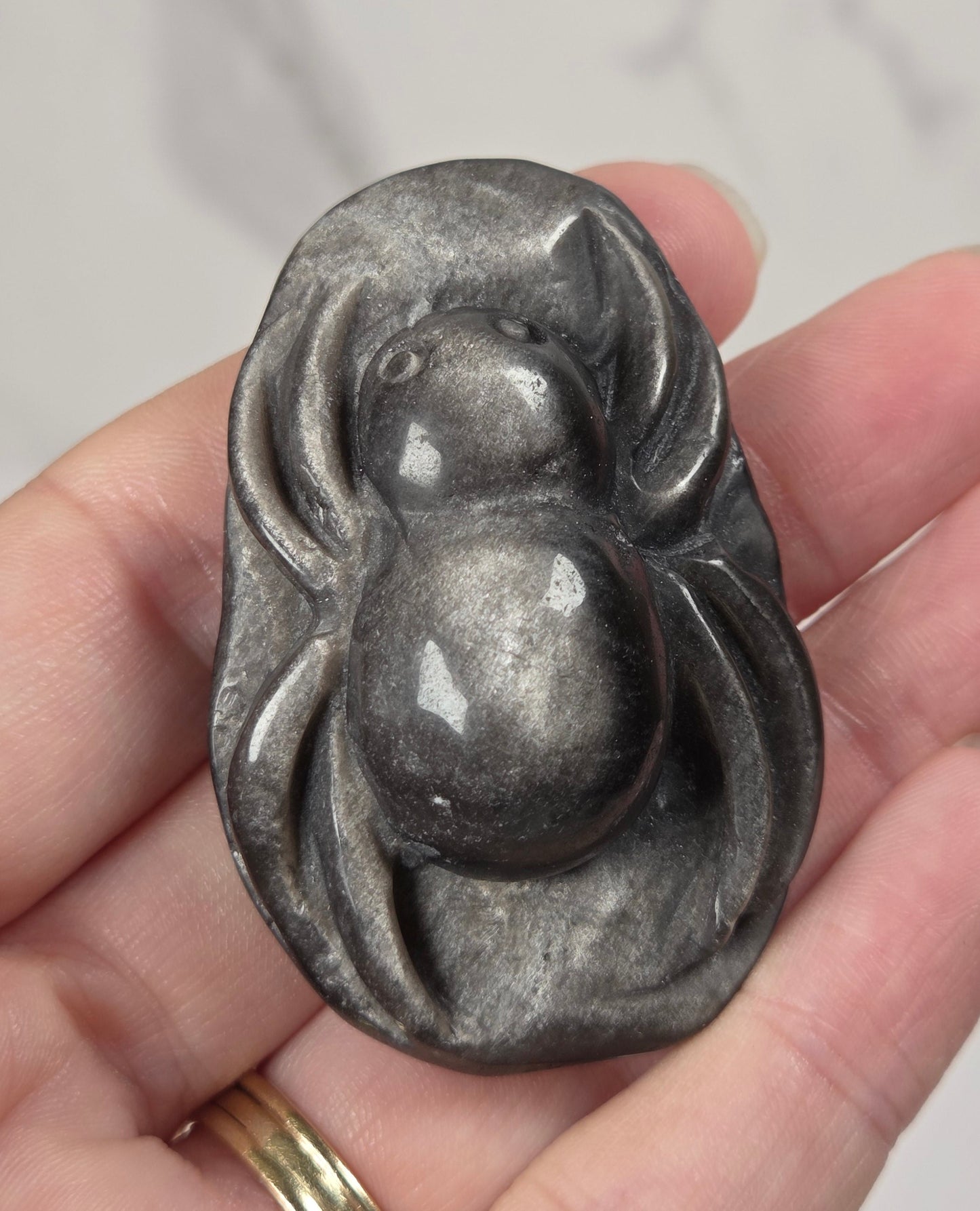 A hand holding a polished dark gray stone pendant shaped like a stylized spider, with a gold ring on one finger, against a light marble background.