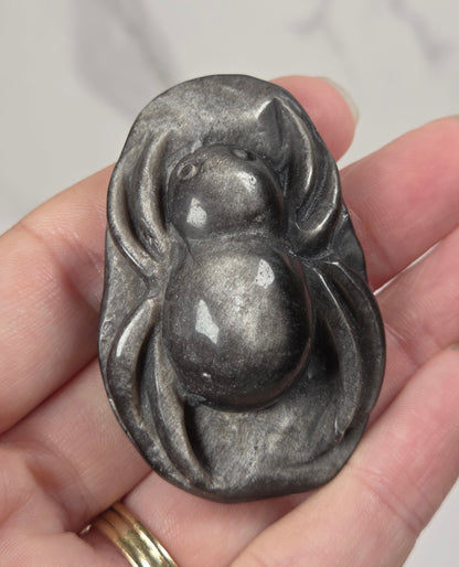 A hand holding a polished dark gray stone pendant shaped like a stylized spider, with a gold ring on one finger, against a light marble background.