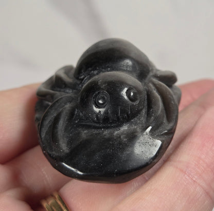 A hand holding a small carved black stone spider figurine, with a gold ring on one finger, against a light gray marble-patterned background.