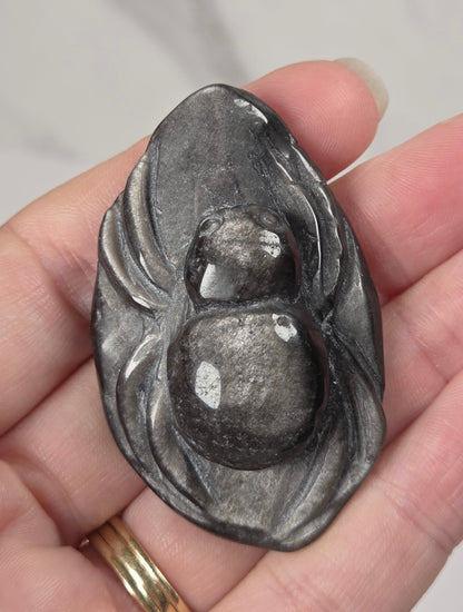 A hand holding a small carved gray stone pendant shaped like a gourd or lotus with a smooth central sphere, against a light marble-like background.