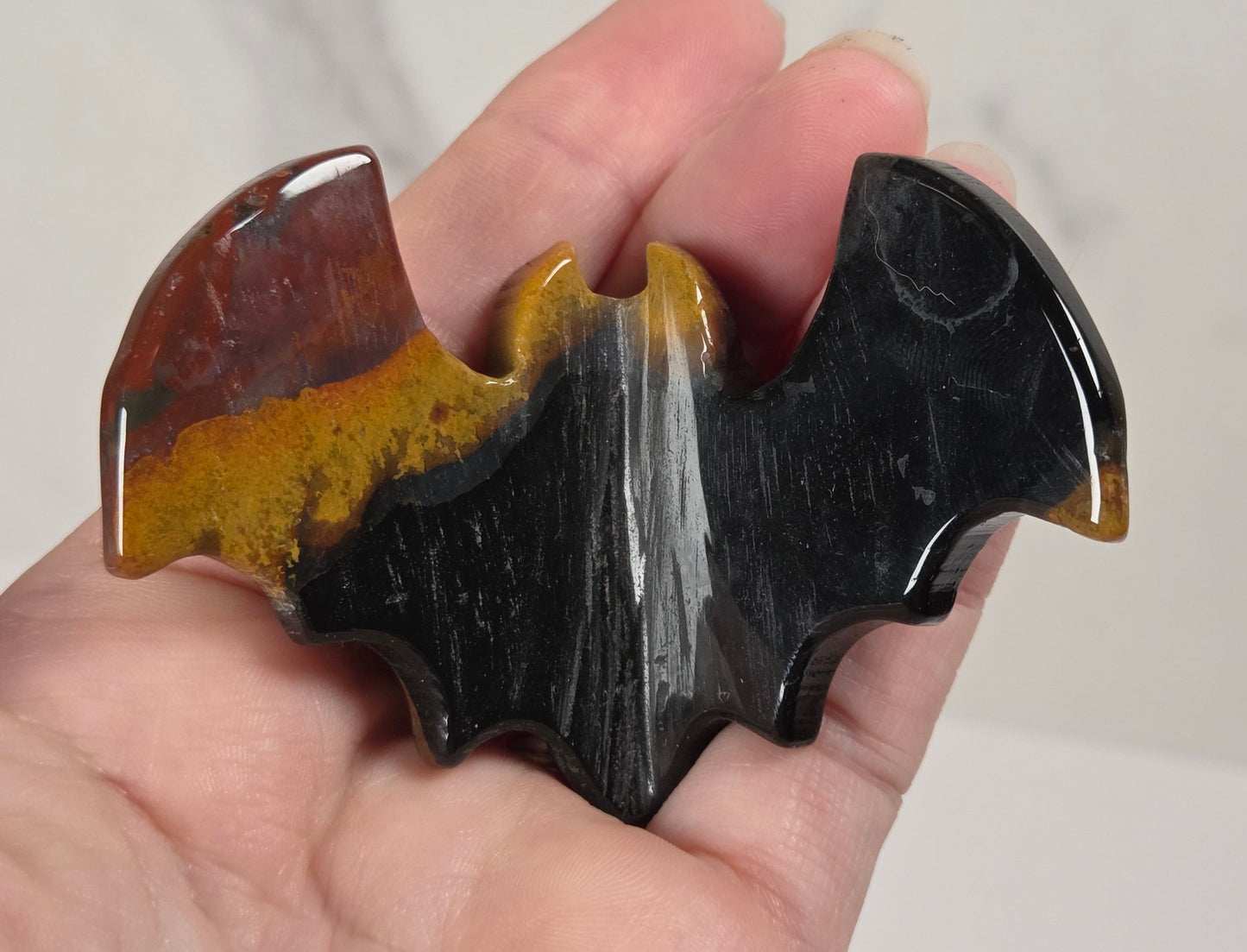 A hand holding a glossy bat-shaped hair claw clip made from a marbled stone with black, brown, and amber-yellow sections, set against a light gray marble background.