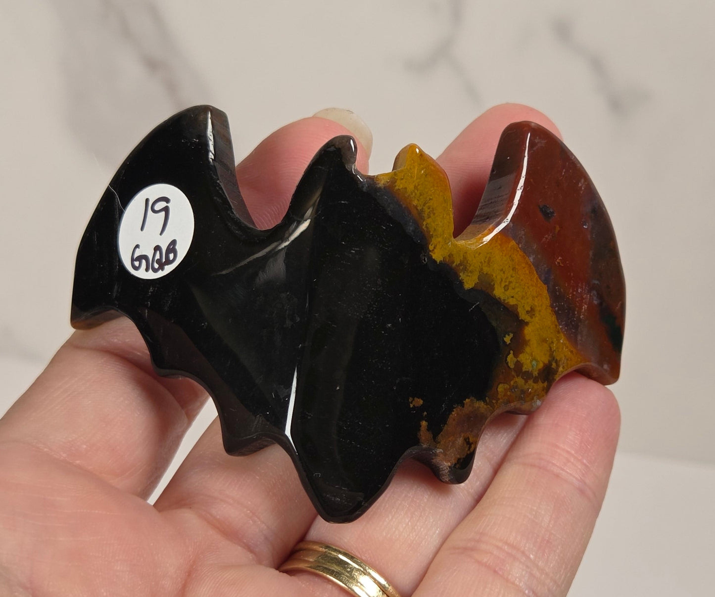 A hand holds a small carved stone object shaped like a bat or winged form. The object has a glossy black section on the left, a reddish-brown section on the right, and a yellow-orange marbled section in the center.