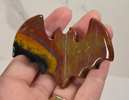 A hand holding a carved bat-shaped pendant made of polished brown and amber stone with a black and yellow band near one wing, against a light marble-patterned background.