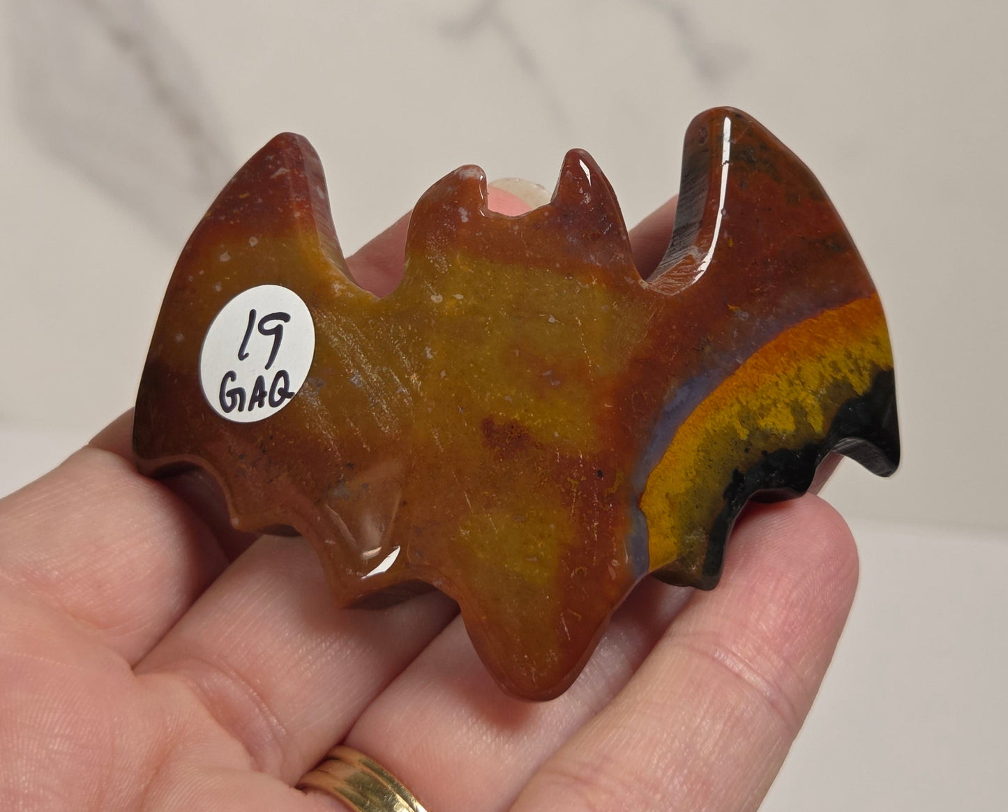 A hand holds a small bat-shaped carved stone or crystal with a mottled brown, amber, and yellow coloration, featuring a small white sticker with handwritten text on the left side.