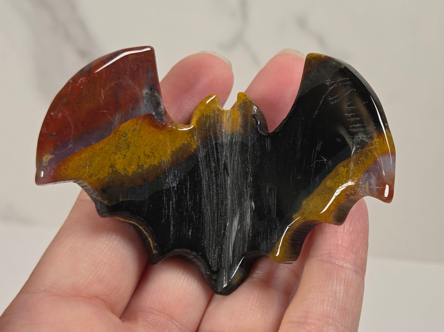 A hand holding a glossy bat-shaped decorative object with a marbled pattern of black, brown, and amber-yellow tones, set against a light marble-patterned background.