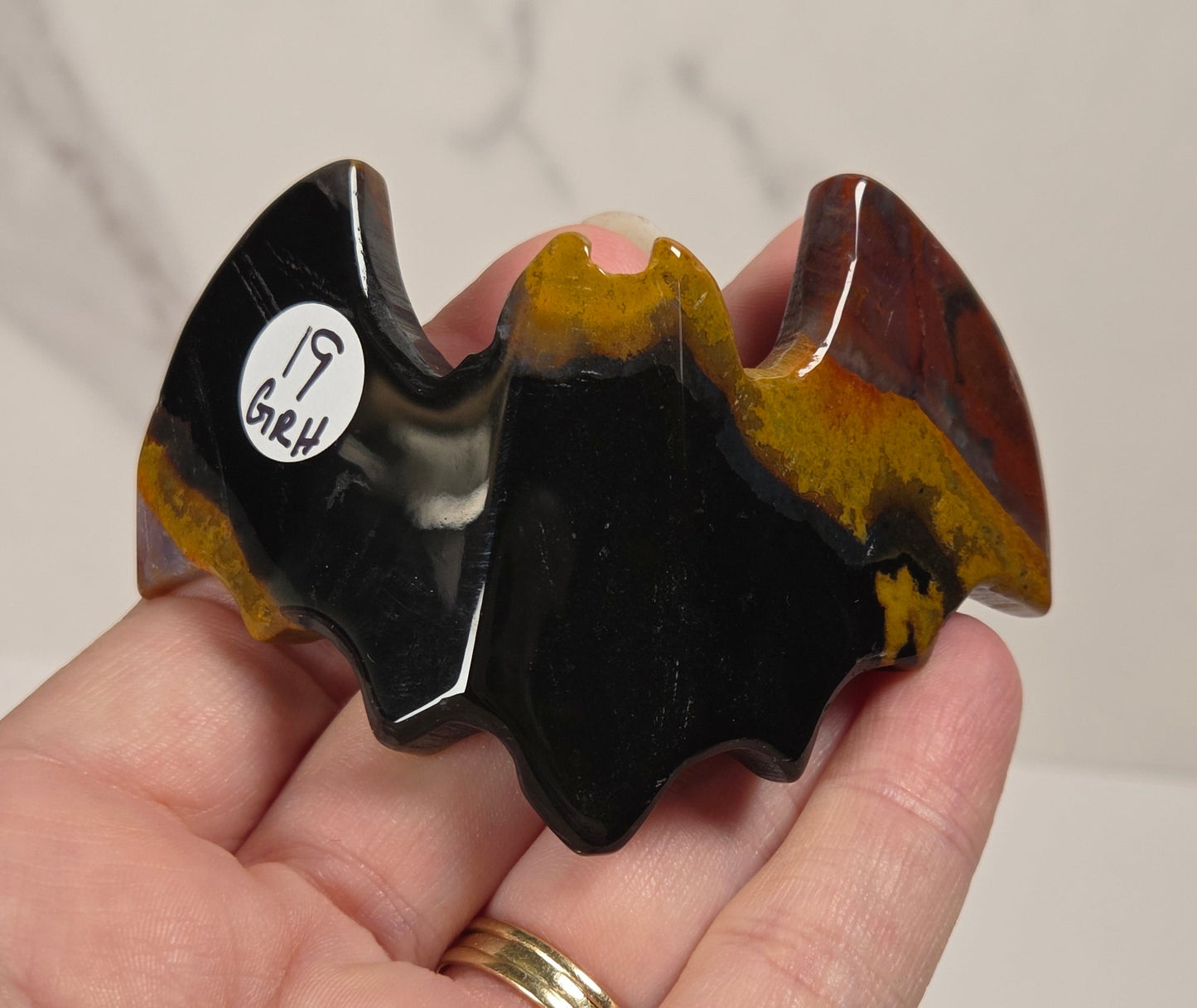 A hand holding a small bat-shaped polished stone object with a black center, yellow and brown marbled wings, and a small white sticker on the side, set against a light marble-patterned background.