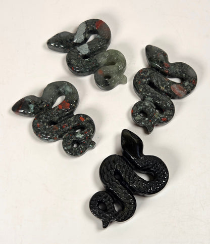Four small snake-shaped carvings  made of speckled dark stone with red inclusions, arranged on a plain off-white background.