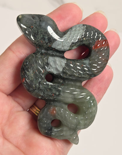 A carved snake-shaped pendant or charm made of polished dark gray stone with subtle red inclusions, held in a person's hand against a light neutral background.