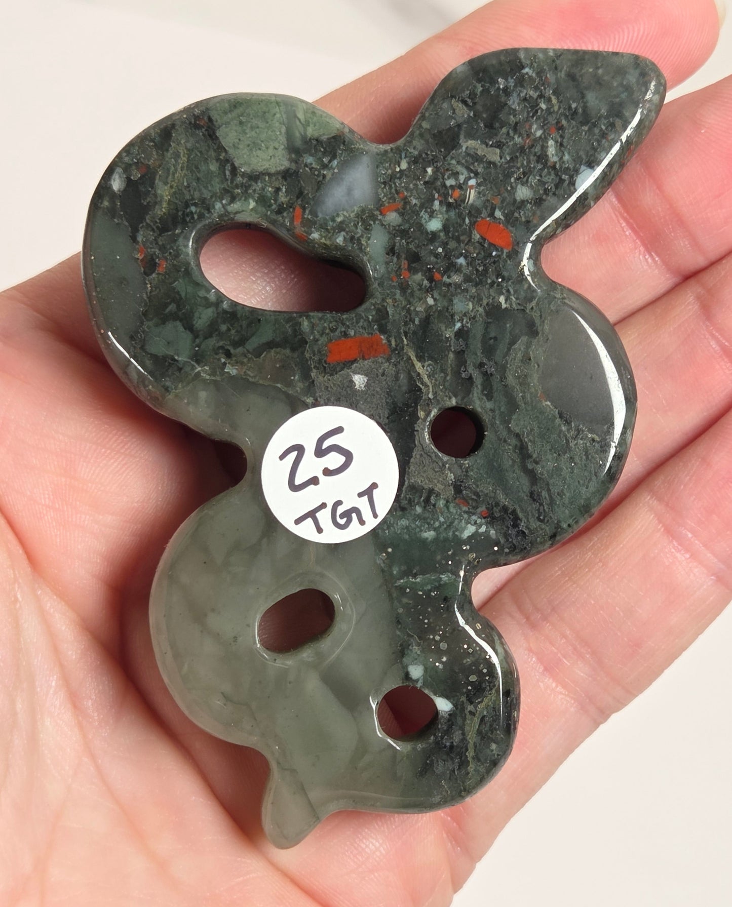 A hand holding a carved green stone pendant with a rabbit-like silhouette, featuring cutouts and a small white sticker that reads '25 TGT'.