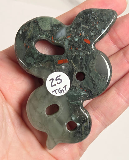 A hand holding a carved green stone pendant with a rabbit-like silhouette, featuring cutouts and a small white sticker that reads '25 TGT'.