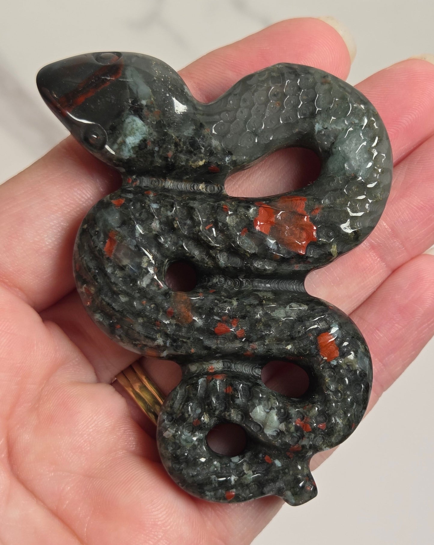 A carved snake-shaped pendant or charm with a textured scale pattern, held in a person's hand. The pendant is primarily dark gray with prominent orange-red and light green stone inclusions, set against a light marble background.