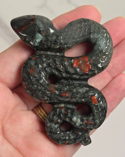 A carved snake-shaped pendant or charm with a textured scale pattern, held in a person's hand. The pendant is primarily dark gray with prominent orange-red and light green stone inclusions, set against a light marble background.
