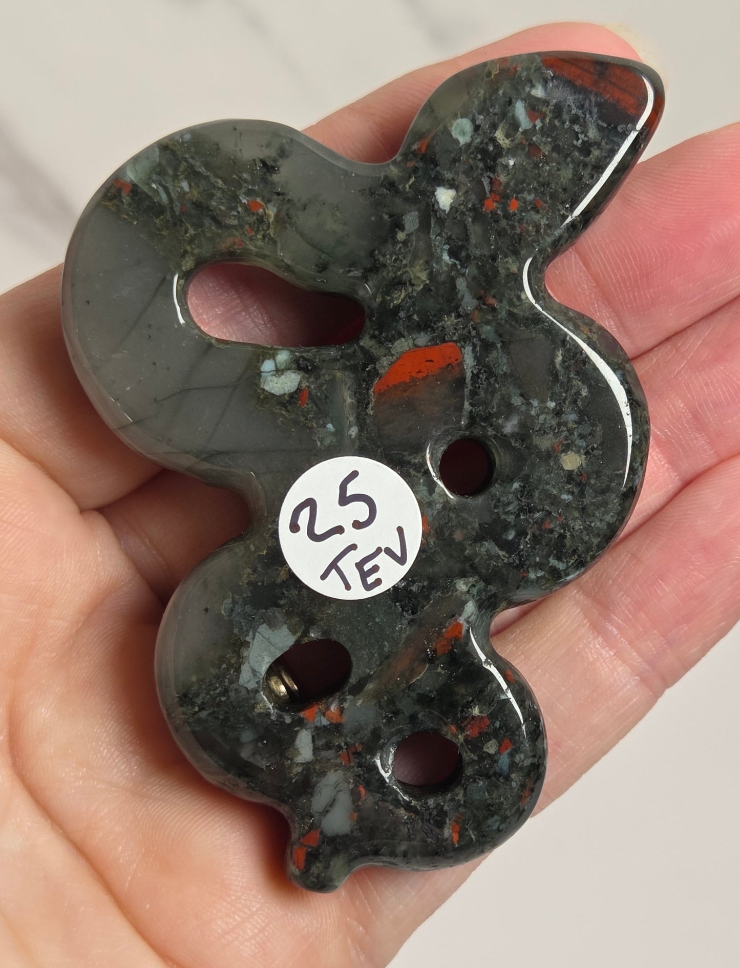 A hand holds a polished dark green stone pendant with orange and white inclusions, featuring three cutouts and a small white sticker reading '25 TEV'.