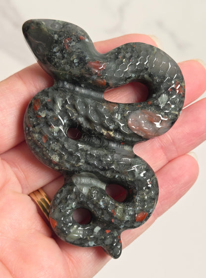A carved snake-shaped pendant or charm made of dark gray stone with red and white inclusions, held in a person's hand against a light neutral background.