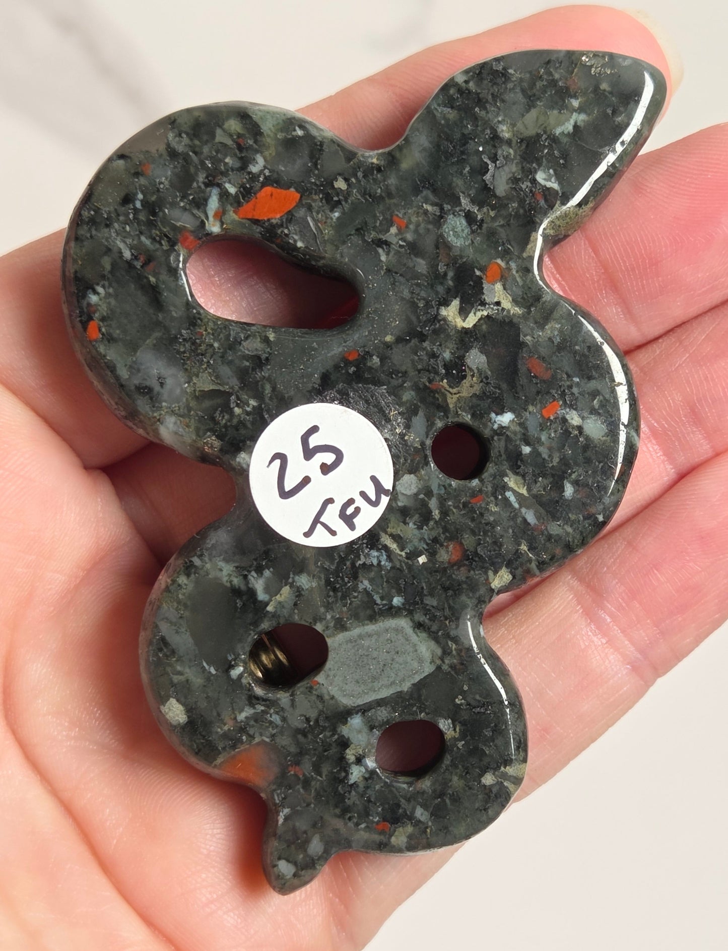 A hand holding a polished dark gray stone charm with orange flecks and a small white sticker that reads '25 KFU'.
