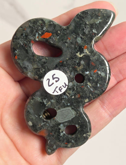 A hand holding a polished dark gray stone charm with orange flecks and a small white sticker that reads '25 KFU'.
