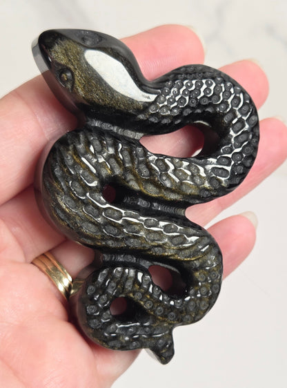 A carved snake figurine or pendant with textured scales, held in a person's hand against a light marble background.
