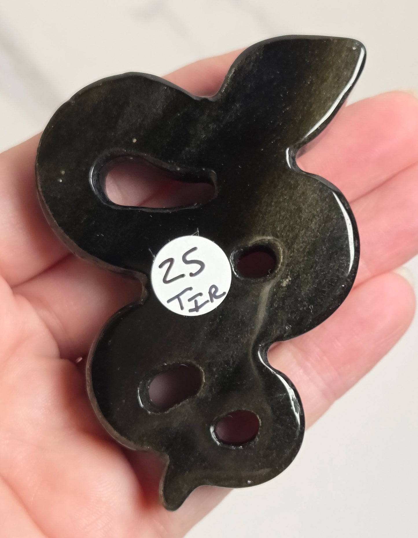 A hand holding a glossy black carved pendant or brooch shaped like a stylized snake or coiled form, with three cutouts and a small white handwritten label reading '25 TIR'.