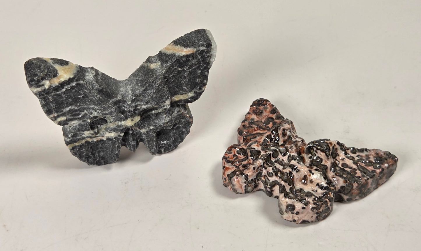 Two small butterfly-shaped stone sculptures on a plain light background. The left butterfly is dark gray with cream veining, and the right butterfly is pinkish beige with black marbling.