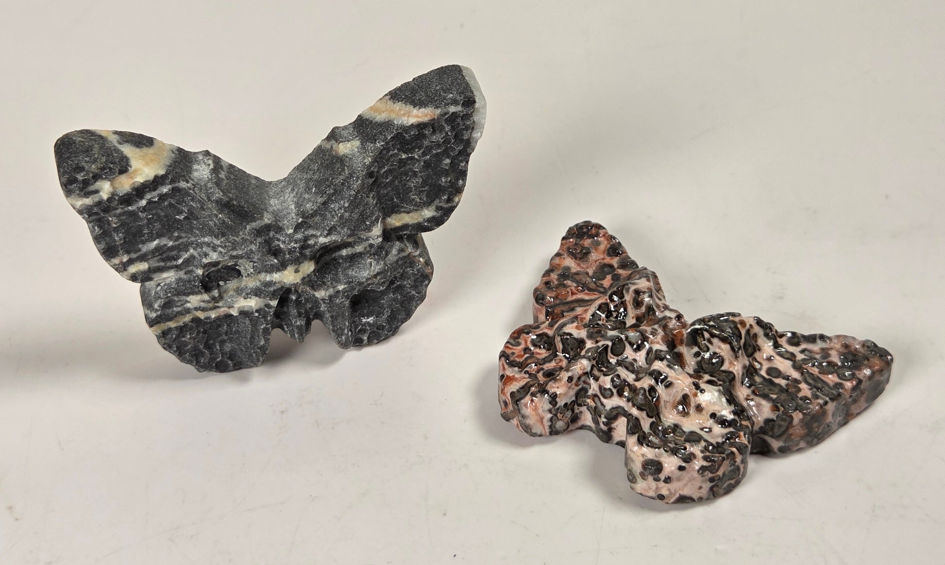 Two small butterfly-shaped stone sculptures on a plain light background. The left butterfly is dark gray with cream veining, and the right butterfly is pinkish beige with black marbling.