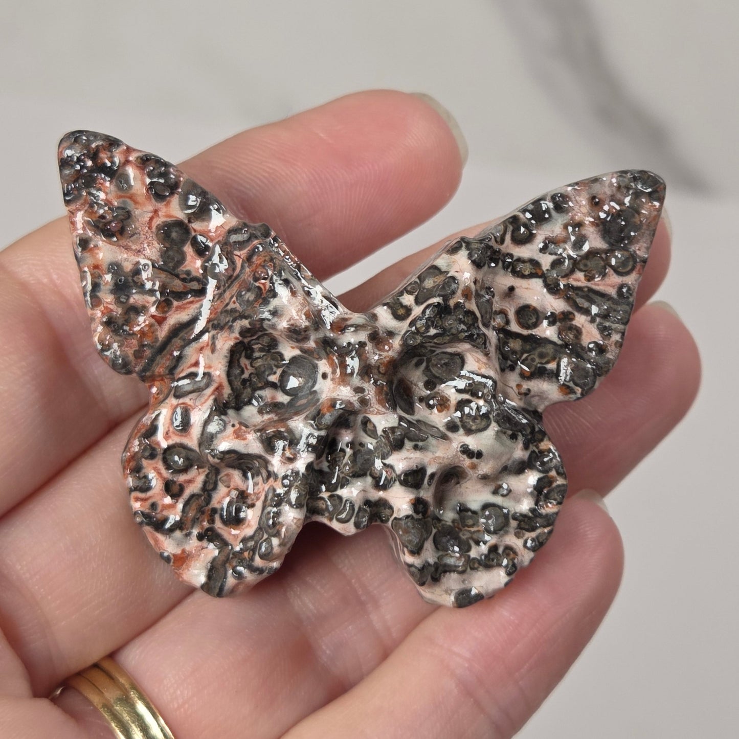 A hand holding a butterfly-shaped brooch or pin with a textured, speckled pattern in black, gray, and warm copper tones over a light beige base.