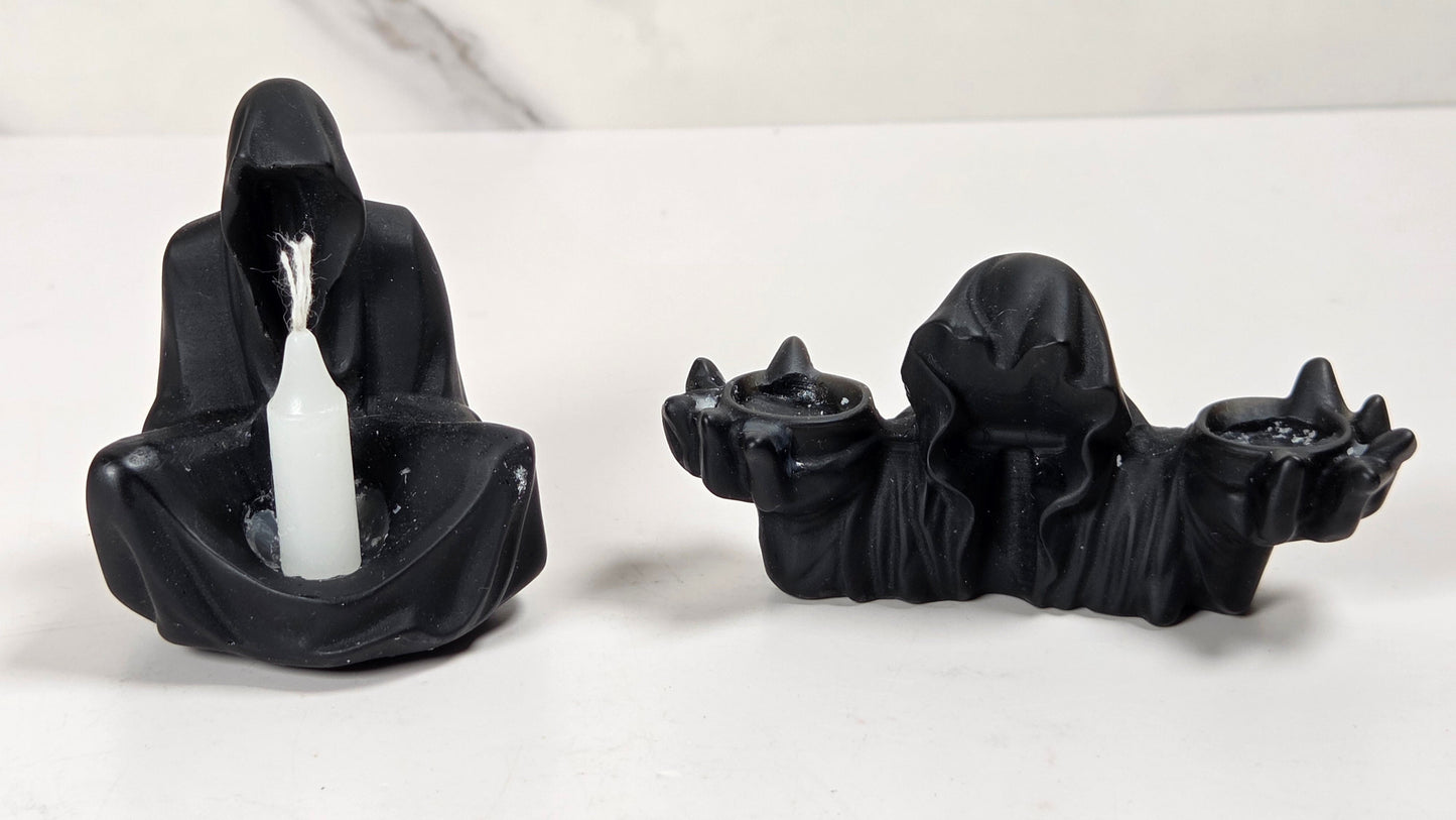 Two black hooded figure candle holders on a white surface with a light gray marble-patterned background. One holder is seated and holds a white taper candle, while the other is elongated and holds two small black candles.