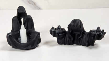 Two black hooded figure candle holders on a white surface with a light gray marble-patterned background. One holder is seated and holds a white taper candle, while the other is elongated and holds two small black candles.