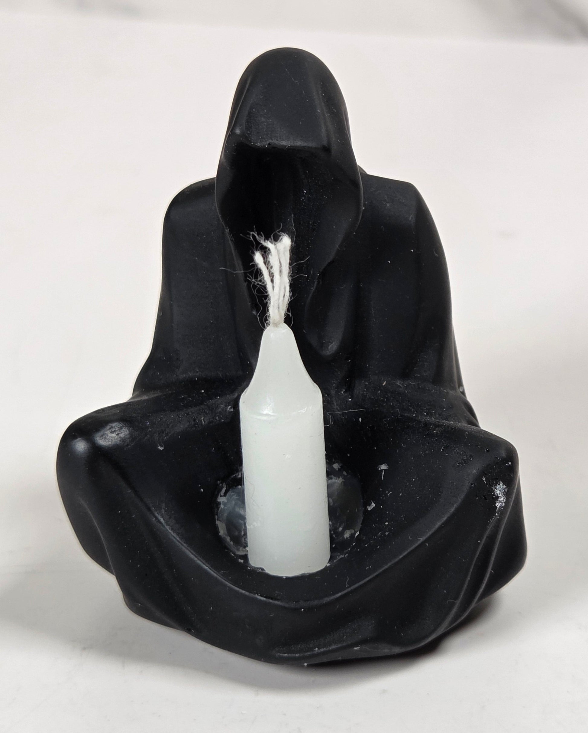A black hooded figure candle holder with a white candle inserted in the center, photographed on a light surface with a marble-patterned background.