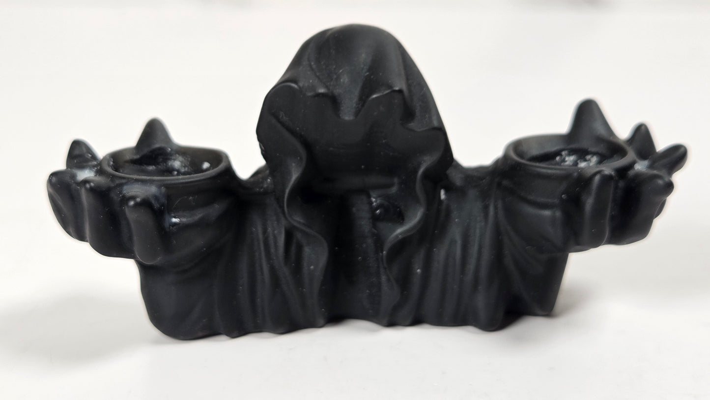 A black gothic-style double candle holder featuring a hooded figure holding two small cups, photographed on a light gray surface.