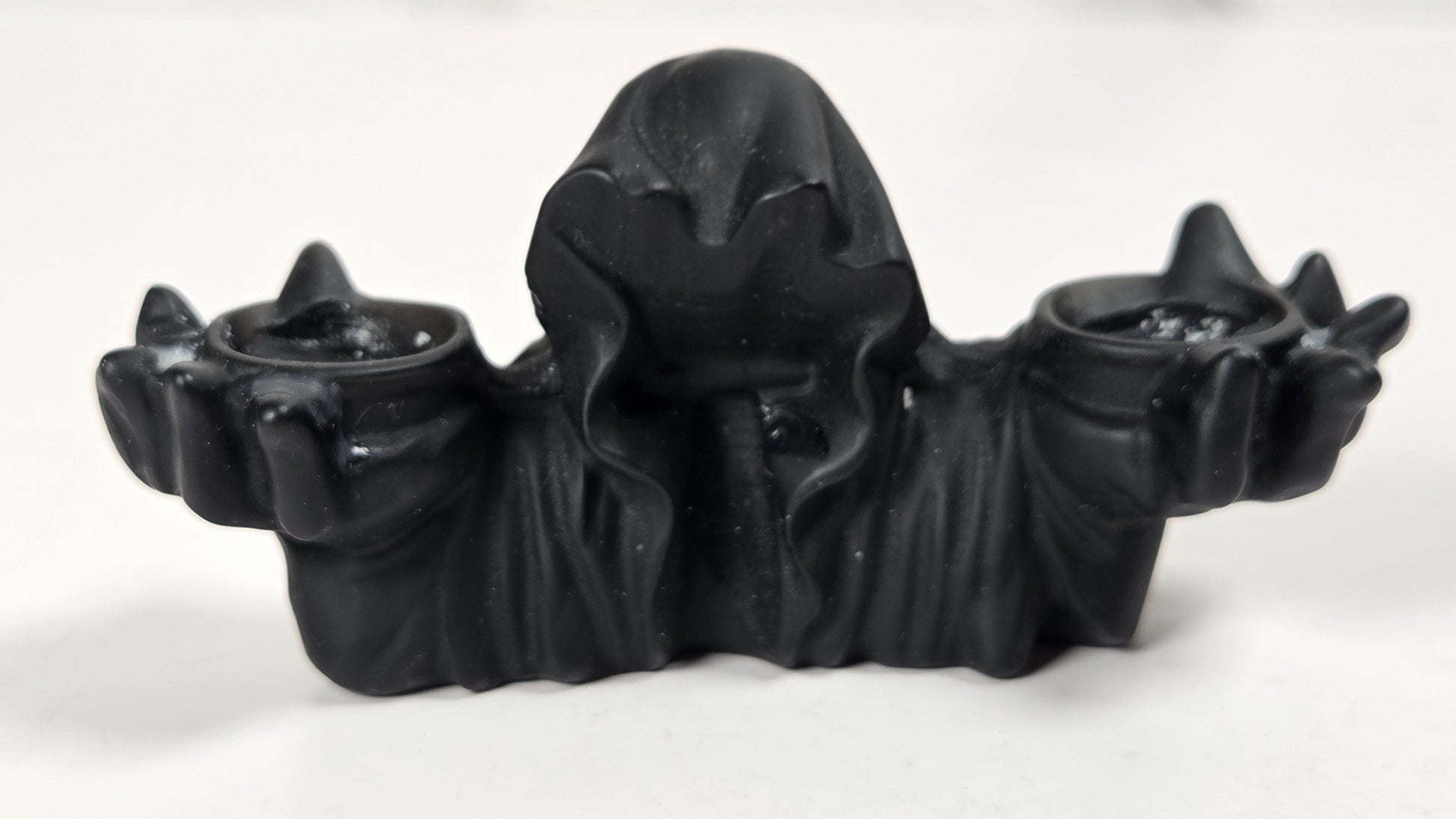 A black gothic-style double candle holder featuring a hooded figure holding two small cups, photographed on a light gray surface.