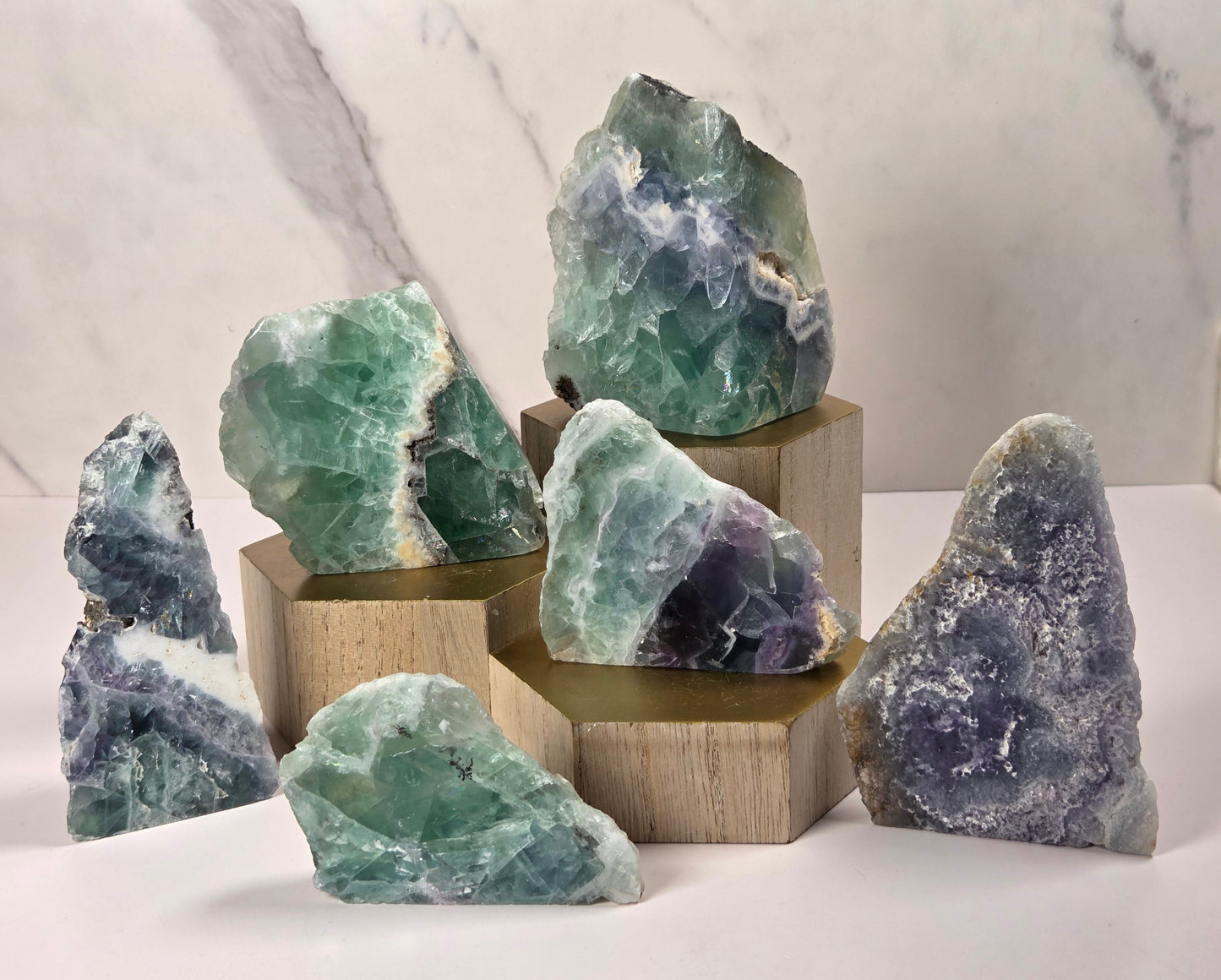 A collection of raw mineral and geode specimens displayed on wooden blocks against a white marble-patterned background. The stones feature natural patterns in shades of green, purple, blue, and gray.
