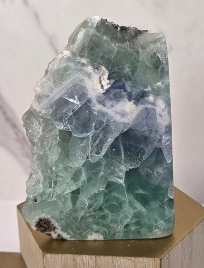 raw mineral and geode specimens displayed on wooden blocks against a white marble-patterned background. The stones feature natural patterns in shades of green, purple, blue, and gray.