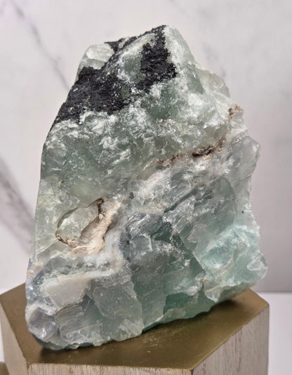 raw mineral and geode specimens displayed on wooden blocks against a white marble-patterned background. The stones feature natural patterns in shades of green, purple, blue, and gray.