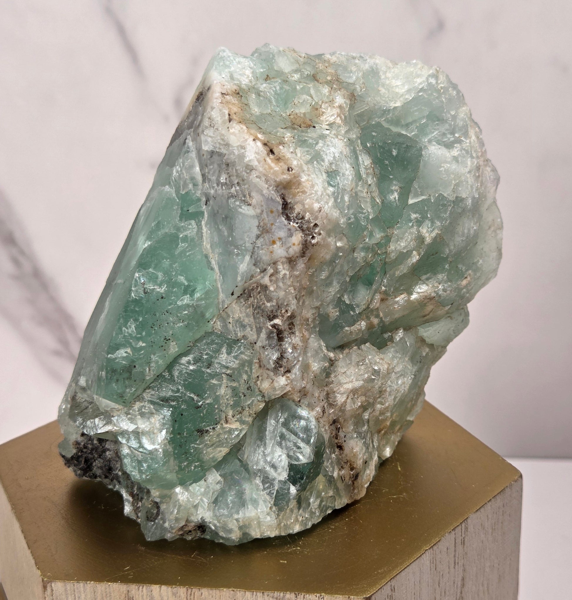A raw green aventurine mineral specimen with natural matrix, displayed on a hexagonal gold-colored wooden pedestal against a light marble-patterned background.
